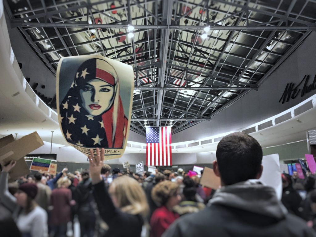 A photo of protesters at the airport holding up "We the people" signs with the American flag in the background.